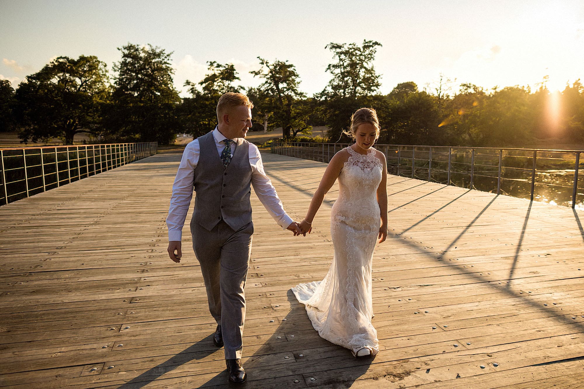 bride and groom in parklands at henham park at golden hour walking hand in hand across the iconic bridge