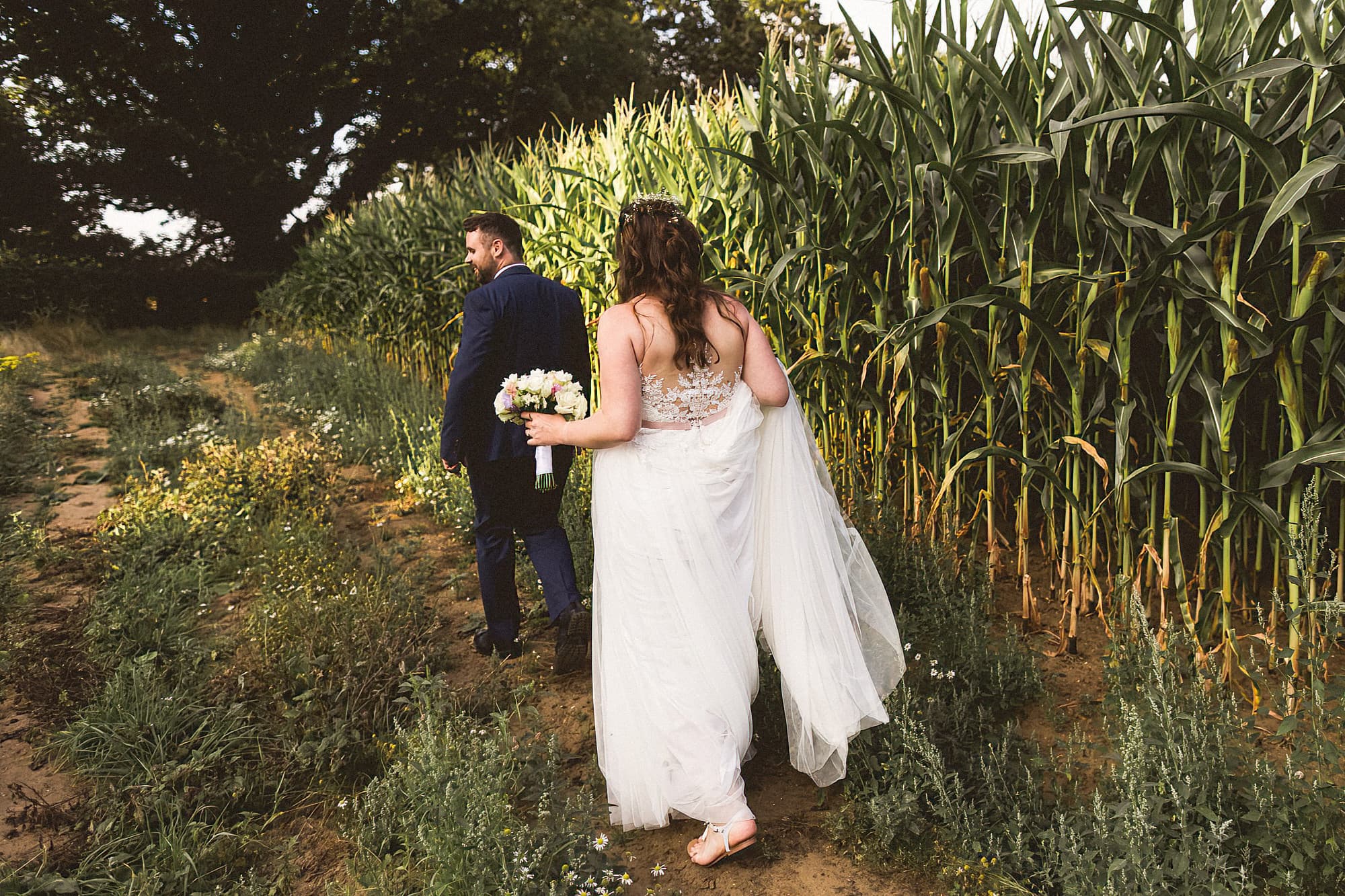 bride and groom walking across field in front of maize