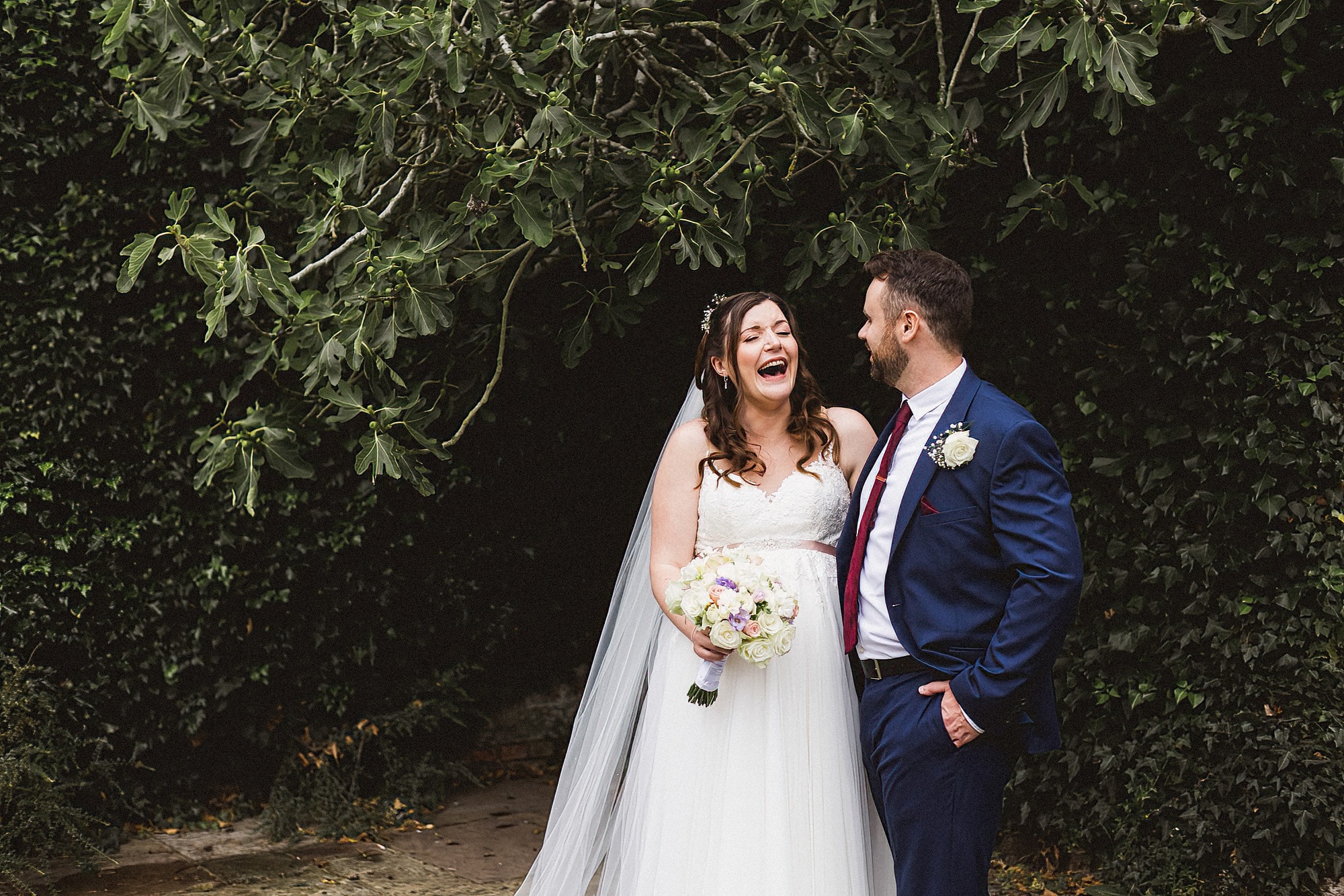bride and groom standing in front of shrubs laughing