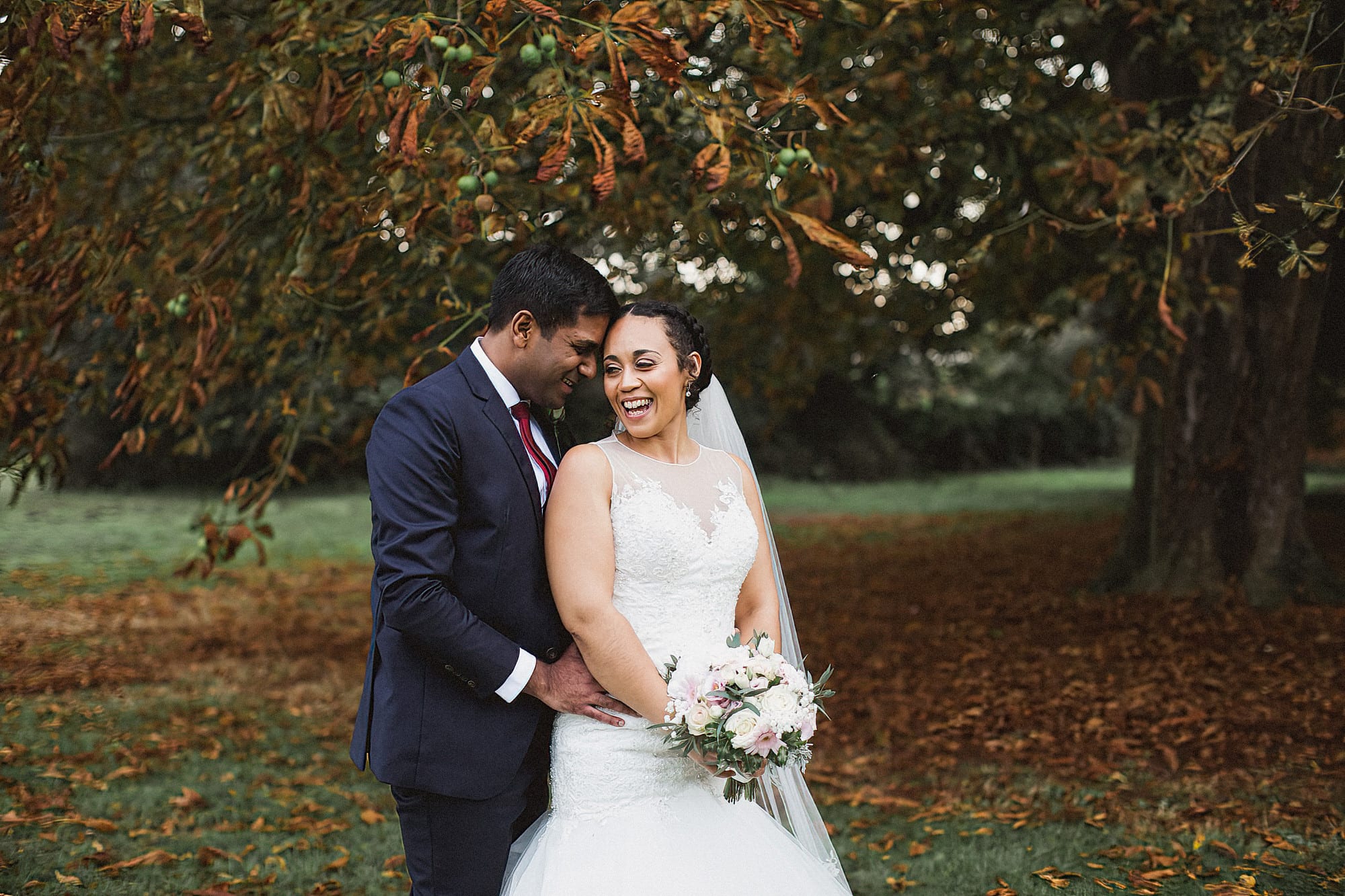 bride and groom standing under tree with rich autumn leaves laughing